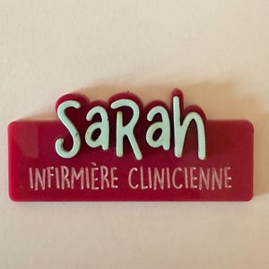 May include: A red name tag with the name "Sarah" in light blue, and the text "INFIRMIERE CLINICIENNE" in white. The name tag is rectangular and has a glossy finish. The name is in a playful, rounded font.