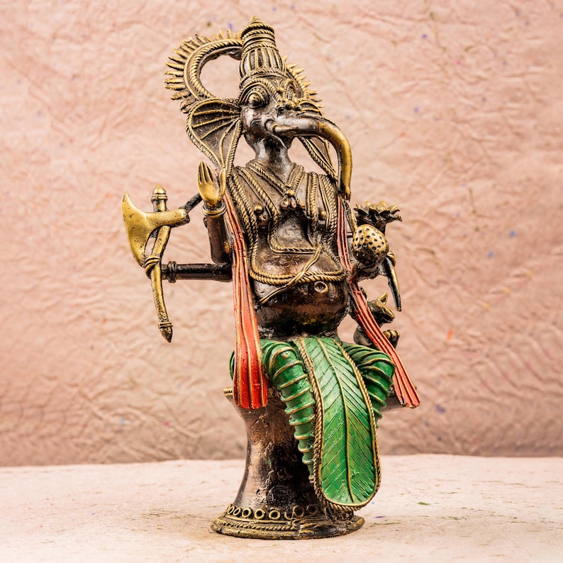 May include: A gold, green, and red metal statue of the Hindu deity Ganesha. The statue depicts Ganesha sitting on a throne with an axe in one hand and a sweet in the other.