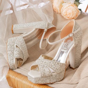 May include: A pair of elegant, open-toe platform heels. The shoes are covered in small, shimmering beads and feature a thick heel and ankle strap. The heels are white and are set on a beige surface with flowers in the background.