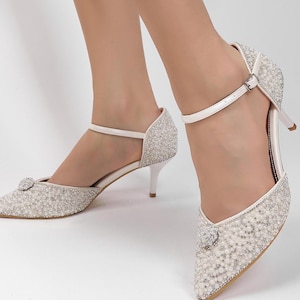 May include: Ivory-colored pointed-toe heels with a delicate ankle strap and a small, stiletto heel. The shoes are embellished with small, sparkling beads and a decorative jewel on the toe. The heels are approximately 2.5 inches high.