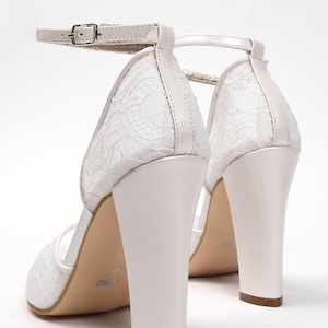 May include: White bridal heels with a block heel and ankle strap. The shoes feature a delicate lace design on the upper and a buckle closure. The heels are approximately 3 inches (7.6 cm) high, perfect for a wedding or special occasion.