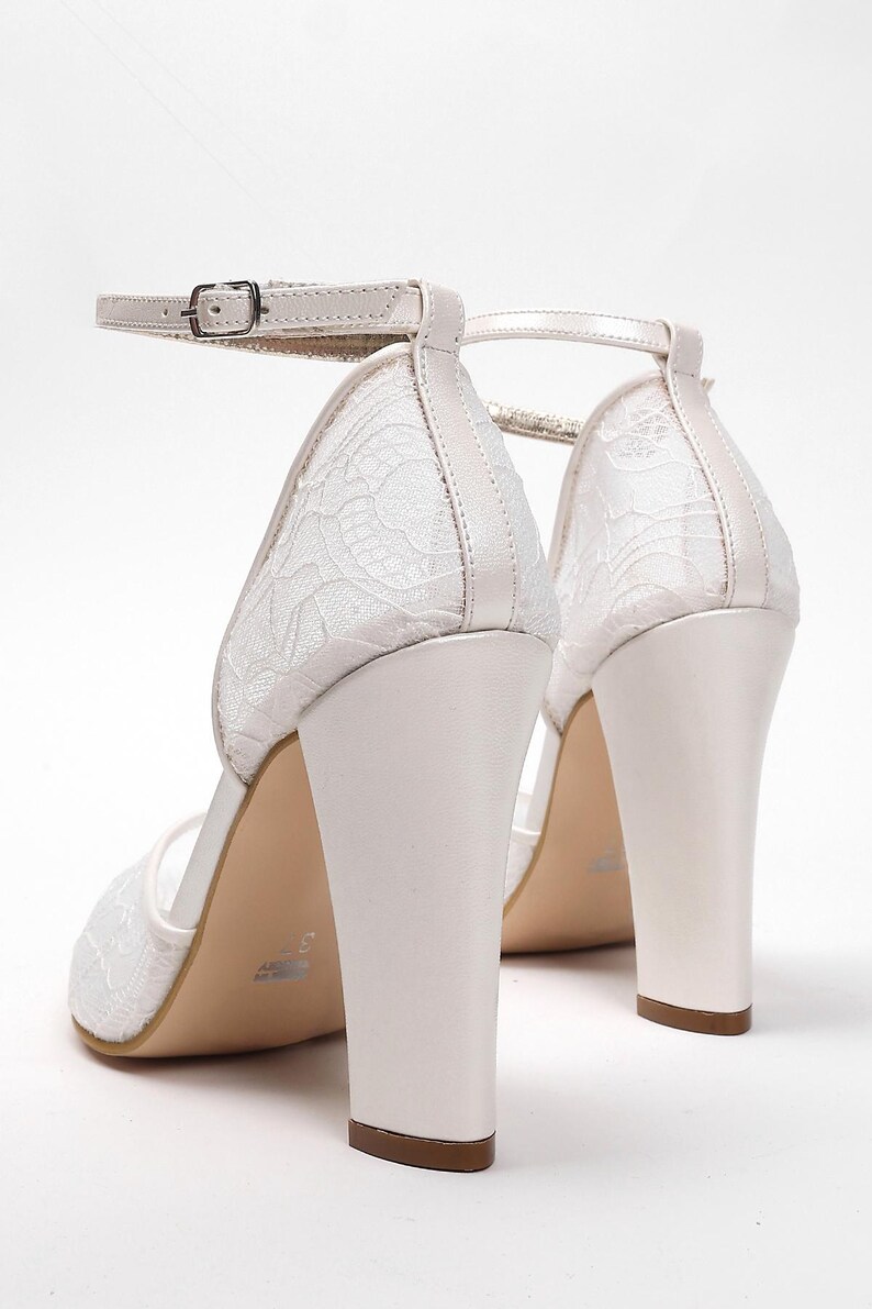 May include: White bridal heels with a block heel and ankle strap. The shoes feature a delicate lace design on the upper and a buckle closure. The heels are approximately 3 inches (7.6 cm) high, perfect for a wedding or special occasion.