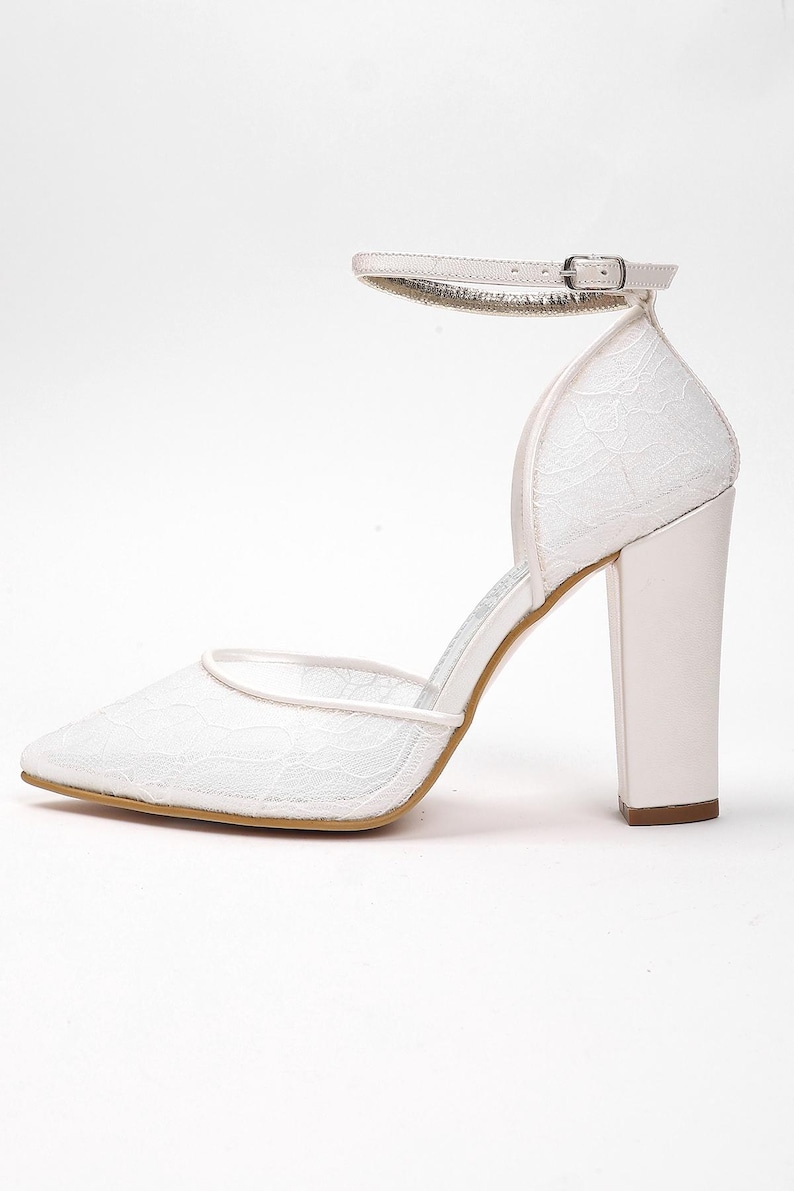 May include: White lace high-heeled shoe with a pointed toe and ankle strap. The heel is a thick, block style. The shoe features a delicate lace pattern and a small buckle on the ankle strap. The heel height is approximately 3.5 inches.