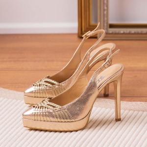 May include: A pair of gold-colored slingback high heels. The shoes have a platform sole and a thin stiletto heel. The toe box features a woven design. The shoes are displayed on a cream-colored rug and a wooden floor.