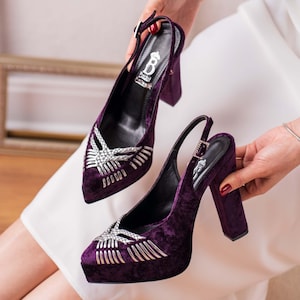 May include: A pair of purple velvet heeled shoes with silver embellishments and a slingback strap. The shoes have a pointed toe and a thick block heel.