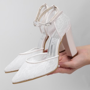 May include: White lace-covered high-heeled shoes with pointed toes and ankle straps. The heels are a pale pink color. The shoes are held up by a hand.