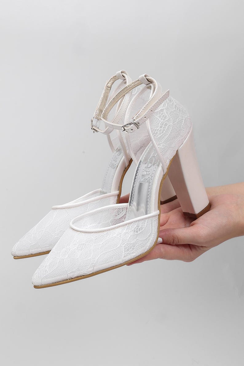 May include: White lace-covered high-heeled shoes with pointed toes and ankle straps. The heels are a pale pink color. The shoes are held up by a hand.