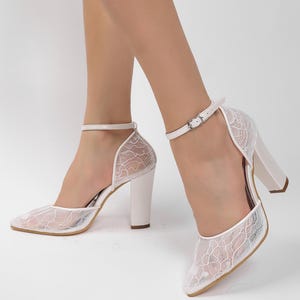 May include: White lace heels with a pointed toe and a thick, block heel. The shoes feature an ankle strap with a silver buckle. The lace overlay creates a delicate, textured appearance, perfect for special occasions.