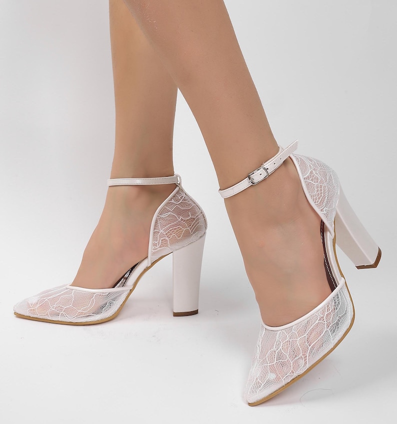 May include: White lace heels with a pointed toe and a thick, block heel. The shoes feature an ankle strap with a silver buckle. The lace overlay creates a delicate, textured appearance, perfect for special occasions.