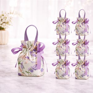 May include: A collection of small, floral-patterned gift bags. The bags feature a cream background with purple and pink floral designs. Each bag has a purple handle and is tied with a purple ribbon. The bags are arranged on a light-colored surface.