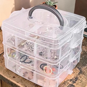 May include: A clear, three-tiered jewelry organizer with a gray handle. The organizer is filled with various jewelry pieces, including necklaces, bracelets, and rings. The clear plastic allows for easy viewing of the contents.