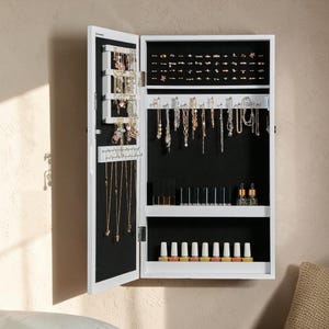 May include: White jewelry armoire with an open door, revealing storage for necklaces, earrings, rings, and other accessories. The interior is black, with shelves and hooks for organizing jewelry and beauty products. The armoire is mounted on a wall.