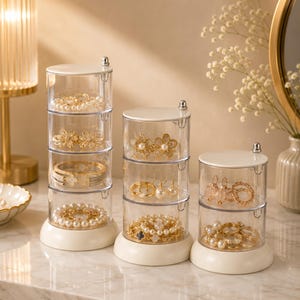 May include: Three clear, stackable jewelry organizers with ivory-colored bases and lids. Each section holds necklaces, bracelets, earrings, and rings. The jewelry includes pearls, gold-toned floral designs, and other decorative pieces.