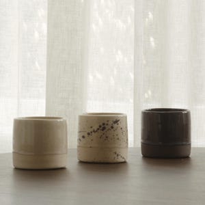 Op de afbeelding: Drie cilindervormige keramische bekers op een rij. De eerste beker is effen lichtcrème van kleur. De tweede beker is crème met donkerbruine verfspatten. De derde beker is effen donkerbruin. Elke beker heeft een gladde, glanzende afwerking.