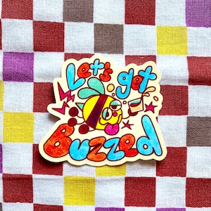 May include: A colorful sticker with the words "Let's Get Buzzed" in blue, orange, and red. The sticker features a cartoon bee with a yellow body, red and black stripes, and a pink tongue. The background is a checkered pattern.