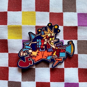 以下が含まれることがあります： 大きな笑顔でメガホンを持っている漫画のキャラクターが特徴のカラフルなエナメルピン。ピンは青い輪郭で、赤、白、紫のチェック柄の背景に配置されています。