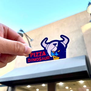 Op de afbeelding: Een gestanste sticker met een gestileerde afbeelding van een gehoornd wezen met een blauw gezicht en een gele kaak. De tekst PIZZA DINOSAUR is gedrukt in dikke rode letters op een donkerblauwe achtergrond. Het ontwerp heeft strakke lijnen.
