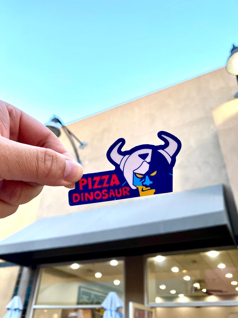 Op de afbeelding: Een gestanste sticker met een gestileerde afbeelding van een gehoornd wezen met een blauw gezicht en een gele kaak. De tekst PIZZA DINOSAUR is gedrukt in dikke rode letters op een donkerblauwe achtergrond. Het ontwerp heeft strakke lijnen.