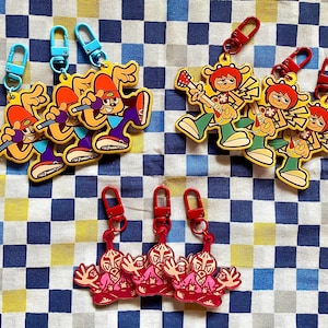 Könnte beinhalten: Schlüsselanhänger mit Cartoon-Charakteren in verschiedenen Posen. Einige Charaktere halten Mikrofone, andere Gitarren, und ein drittes Design ist ebenfalls vorhanden. Jeder Schlüsselanhänger hat einen farbigen Verschluss. Der Hintergrund ist ein kariertes Muster.