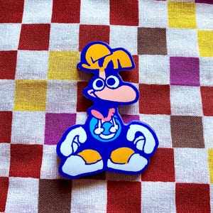 May include: A blue cartoon character sticker with white, orange, and pink accents. The sticker features a character with large eyes, a pink nose, and a yellow headpiece. The character is set against a checkered background.
