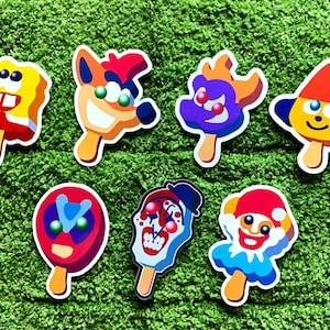 May include: Seven colourful stickers in the shape of ice lollies, each depicting a cartoon character. The designs include a yellow sponge, a fox, a purple creature, a dog, a superhero, a clown, and a smiling clown.