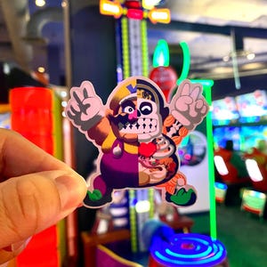 Op de afbeelding: Een gestanste sticker met een gespleten ontwerp van een stripfiguur. De linkerkant toont een geel en paars personage met een snor die een vredesteken maakt, terwijl de rechterkant een anatomisch skelet en interne organen onthult.