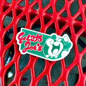 Op de afbeelding: Een gestanste sticker met een groene rechthoekige achtergrond met afgeronde randen. De tekst Gelato Joe's is geschreven in een vetgedrukt, rood, afgerond lettertype. Rechts van de tekst staat een witte grafische illustratie van een gestileerd, lachend stripfiguur.