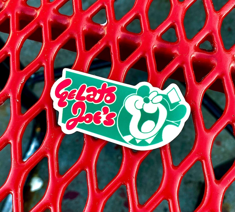 Op de afbeelding: Een gestanste sticker met een groene rechthoekige achtergrond met afgeronde randen. De tekst Gelato Joe's is geschreven in een vetgedrukt, rood, afgerond lettertype. Rechts van de tekst staat een witte grafische illustratie van een gestileerd, lachend stripfiguur.