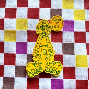 Op de afbeelding: Gele sticker met een cartoonfiguur op rolschaatsen met de tekst "ROCK IT ON" in een tekstballon. De figuur is groen omlijnd, op een geruite achtergrond.
