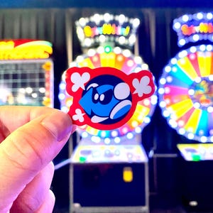Op de afbeelding: Een gestanste sticker met een gestileerd blauw personage met een donkerblauw masker en witte accenten, tegen een levendige rode achtergrond met witte wolkachtige vormen rondom de centrale figuur in een gedurfd grafisch ontwerp.