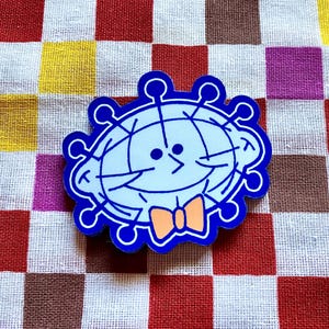 May include: A blue and white cartoon pin with a smiling face, a bow tie, and a spiky outline. The pin is set against a colorful checkered background. The design is whimsical and playful.