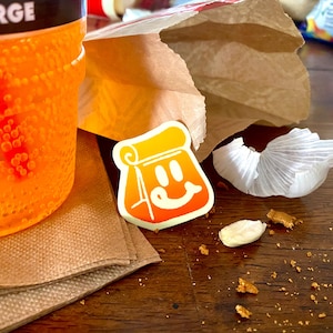 Op de afbeelding: Een feloranje koolzuurhoudende drank in een grote plastic beker naast een bruine papieren zak, een witte sticker met een gestileerd oranje papieren zak-figuurtje met een lachend gezicht, een stukje witte knoflookschil en bruine kruimels.