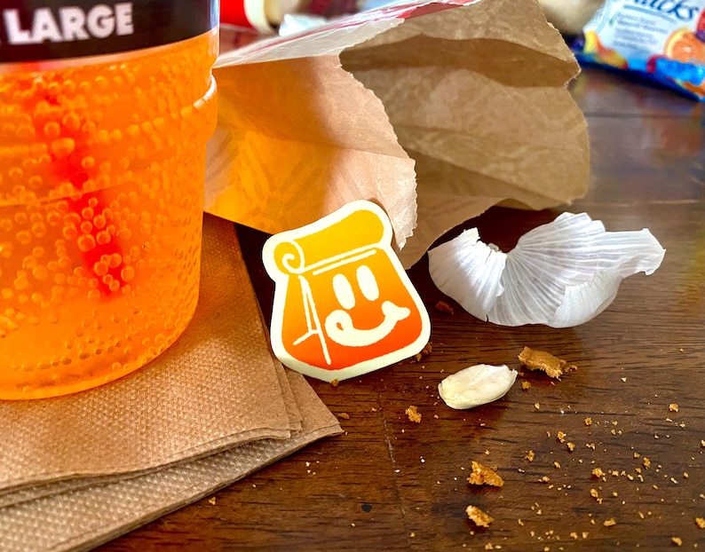 Op de afbeelding: Een feloranje koolzuurhoudende drank in een grote plastic beker naast een bruine papieren zak, een witte sticker met een gestileerd oranje papieren zak-figuurtje met een lachend gezicht, een stukje witte knoflookschil en bruine kruimels.