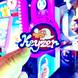 Op de afbeelding: Een gestanste sticker met een stripfiguur met een wit hoofd en rode ogen, die een geel object in de mond houdt. Het woord Keyzer staat in een vetgedrukt, wit, gestileerd lettertype onderaan de sticker tegen een donkerrode cirkel aan.