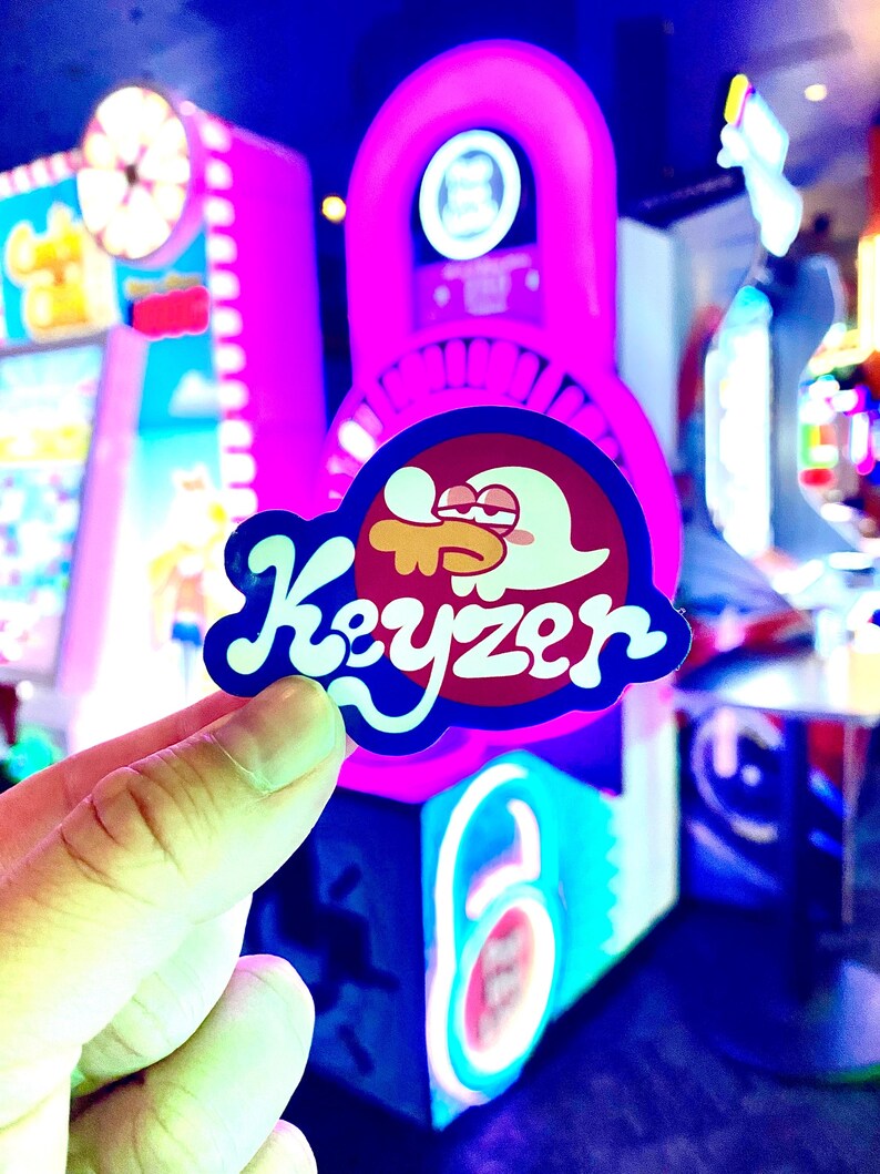 Op de afbeelding: Een gestanste sticker met een stripfiguur met een wit hoofd en rode ogen, die een geel object in de mond houdt. Het woord Keyzer staat in een vetgedrukt, wit, gestileerd lettertype onderaan de sticker tegen een donkerrode cirkel aan.