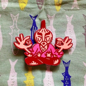 May include: A red and pink enamel pin featuring a seated figure with hands raised, holding dice. The pin is set against a green background with a fish pattern. The figure has a distinctive face and is in a meditative pose.