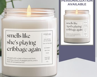 May include: A white candle in a glass jar with a label that reads "smells like she's playing cribbage again". The candle is lit and the flame is visible. The label also includes the text "Infused with a dash of back-and-forth scoring, a touch of pegged points, and a hint of competitive fun."