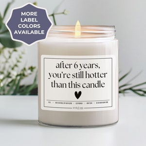 May include: A white candle with a black label that reads "after 6 years, you're still hotter than this candle". The candle is in a clear glass jar and has a small black heart on the label.