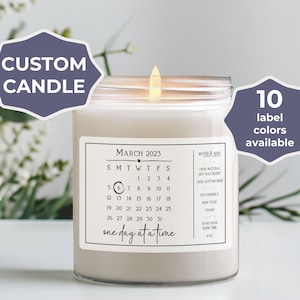 May include: A white candle with a label that says "March 2023" and has a calendar with the number 6 circled. The label also says "one day at a time". The candle is in a clear glass jar. The background is a light gray.