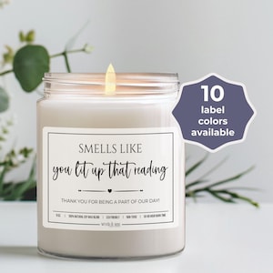 May include: A white candle with a clear glass jar and a label that reads "Smells Like You Lit Up That Reading" with a heart symbol. The label also says "Thank You For Being A Part Of Our Day!" and has a small text at the bottom. The candle is lit and the flame is visible. There is a purple circle with white text that says "10 label colors available".