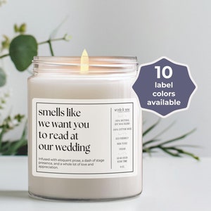 May include: A white candle in a glass jar with a label that reads "smells like we want you to read at our wedding". The label also includes information about the candle, such as the fact that it is soy wax and has a cotton wick. The candle is sitting on a white surface with green plants in the background.