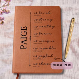 May include: A brown leather journal with the name "PAIGE" vertically printed on the left side. The journal features inspirational text and a gold pen. The text includes "is loved", "is strong", and "is chosen".