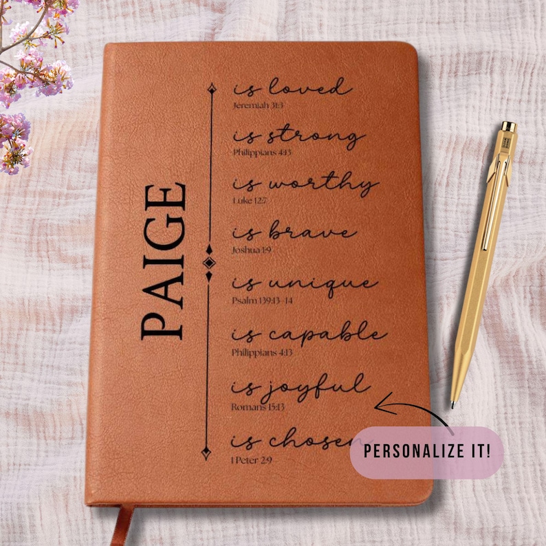 May include: A brown leather journal with the name "PAIGE" vertically printed on the left side. The journal features inspirational text and a gold pen. The text includes "is loved", "is strong", and "is chosen".