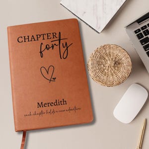 Puede incluir: Un diario de cuero marr&oacute;n con las palabras "CHAPTER forty" y un s&iacute;mbolo de coraz&oacute;n. El nombre "Meredith" y la frase "each chapter holds a new adventure" tambi&eacute;n est&aacute;n impresos en la portada. Un posavasos tejido, un port&aacute;til y un rat&oacute;n est&aacute;n cerca.