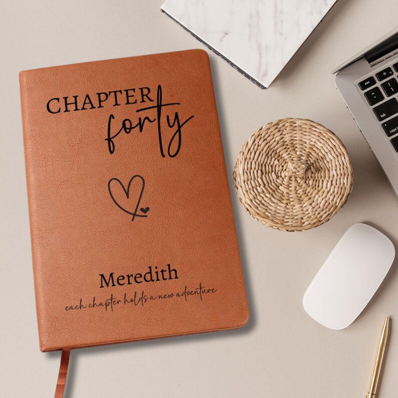 Puede incluir: Un diario de cuero marr&oacute;n con las palabras "CHAPTER forty" y un s&iacute;mbolo de coraz&oacute;n. El nombre "Meredith" y la frase "each chapter holds a new adventure" tambi&eacute;n est&aacute;n impresos en la portada. Un posavasos tejido, un port&aacute;til y un rat&oacute;n est&aacute;n cerca.