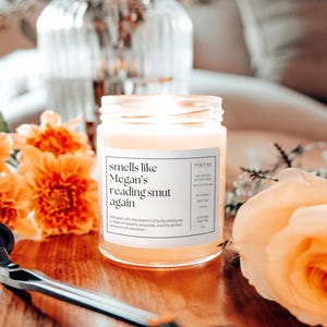 Op de afbeelding: Een glazen pot met een witte sticker met de tekst "smells like Megan's reading smut again". De kaars is aangestoken en de was is lichtgeel. De kaars is omringd door oranje en gele bloemen.
