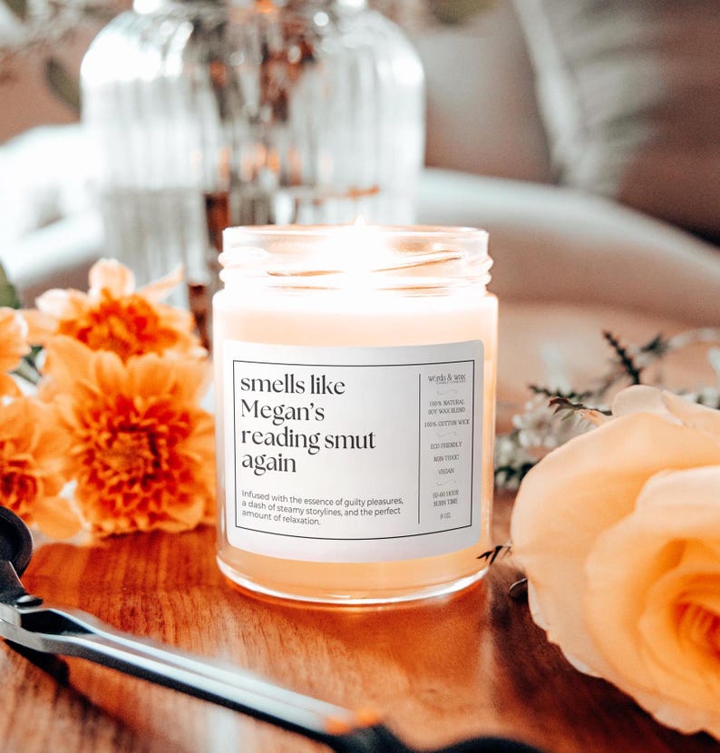 Op de afbeelding: Een glazen pot met een witte sticker met de tekst "smells like Megan's reading smut again". De kaars is aangestoken en de was is lichtgeel. De kaars is omringd door oranje en gele bloemen.