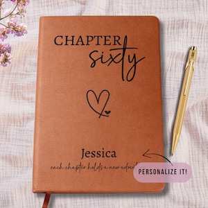 May include: A brown leather-bound journal with the words "CHAPTER sixty" and a heart symbol. The name "Jessica" is below the heart, with the phrase "each chapter holds a new adventure." A gold pen rests beside the journal. The journal is approximately 8 inches tall.