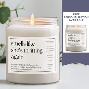 May include: A white candle with a clear glass jar and a black label that reads "smells like she's thrifting again". The label also includes the text "Infused with the thrill of the hunt, a sprinkle of budget-savvy brilliance, and a hint of secondhand style."