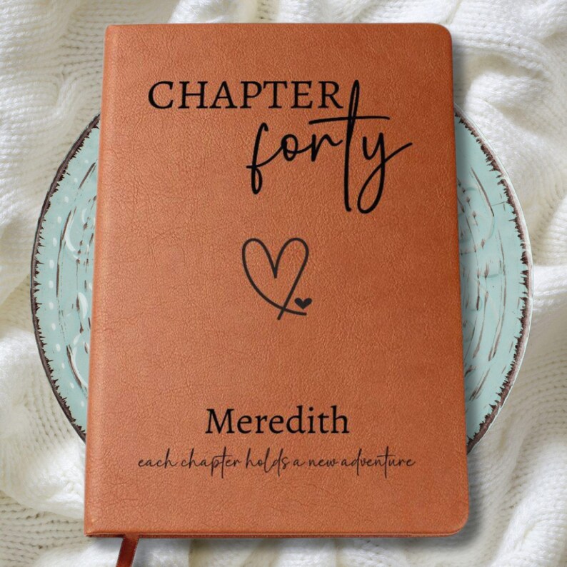 Puede incluir: Un diario de cuero marr&oacute;n con las palabras "CHAPTER forty" y el nombre "Meredith" impresos en la portada. Un gr&aacute;fico de coraz&oacute;n est&aacute; entre las palabras y el nombre. La frase "each chapter holds a new adventure" est&aacute; impresa en la parte inferior.